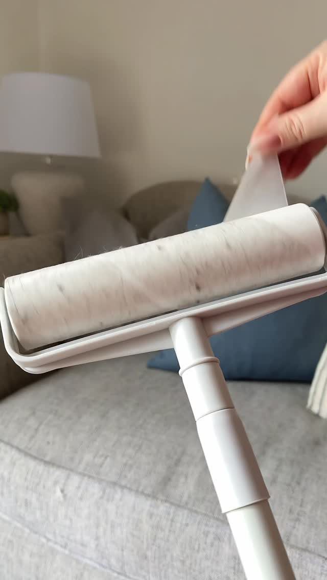 A hand peeling a used sheet from a white lint roller on a grey sofa.