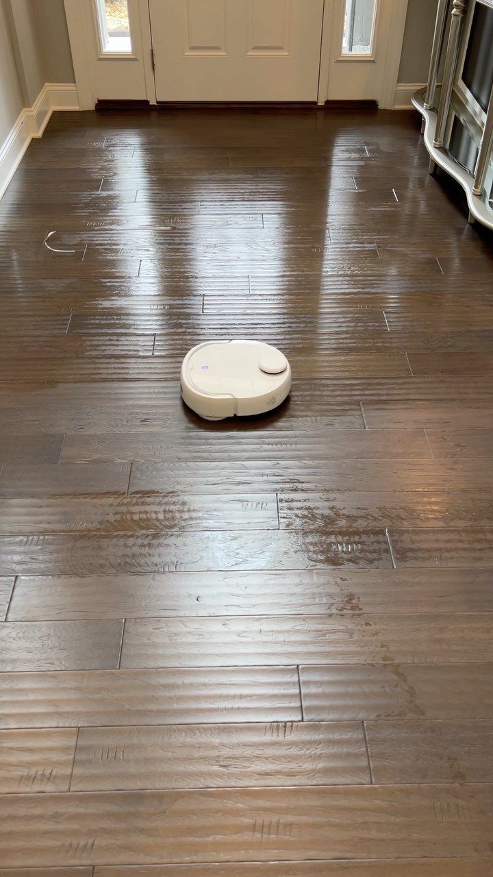A white robotic vacuum cleaner on a dark wood floor in a bright hallway.