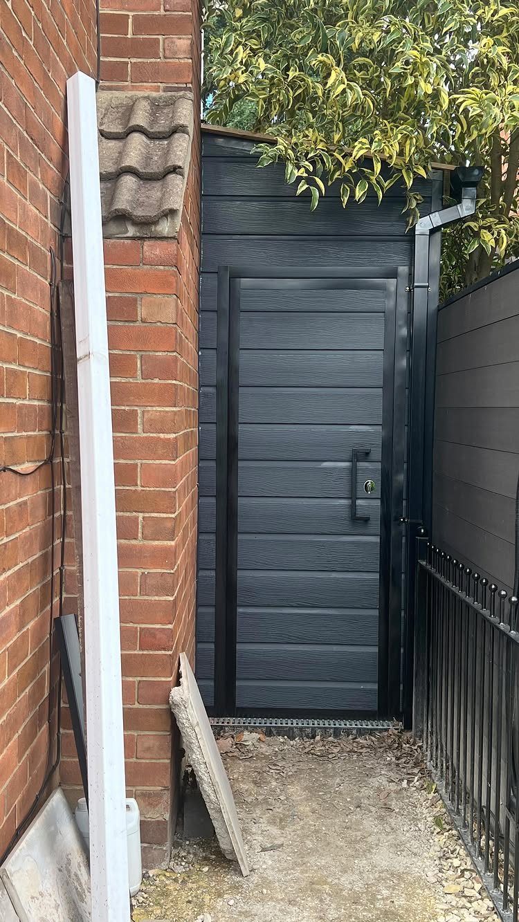 8 Lean-To Shed Ideas for Storage, Parking and Backyard Layouts 4 A dark grey textured lean-to shed positioned against a red brick wall in a narrow outdoor space.