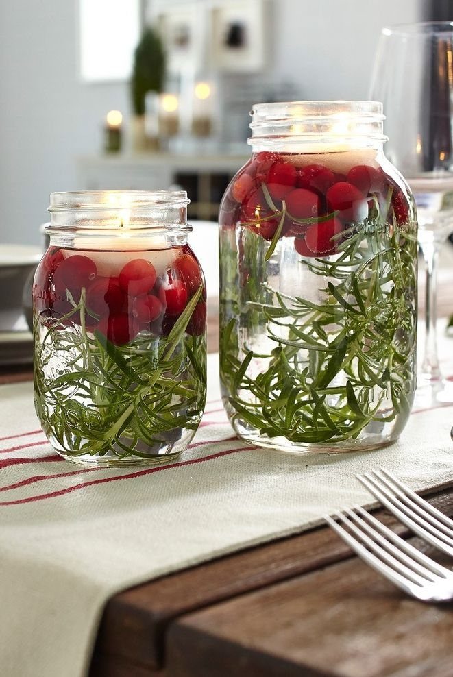 Two mason jars filled with rosemary, red cranberries, and floating lit tea light candles.