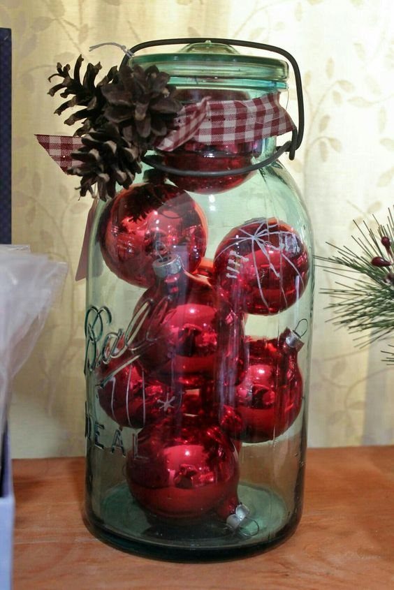 A large glass jar filled with shiny red ornaments, decorated with a pinecone and checkered ribbon.
