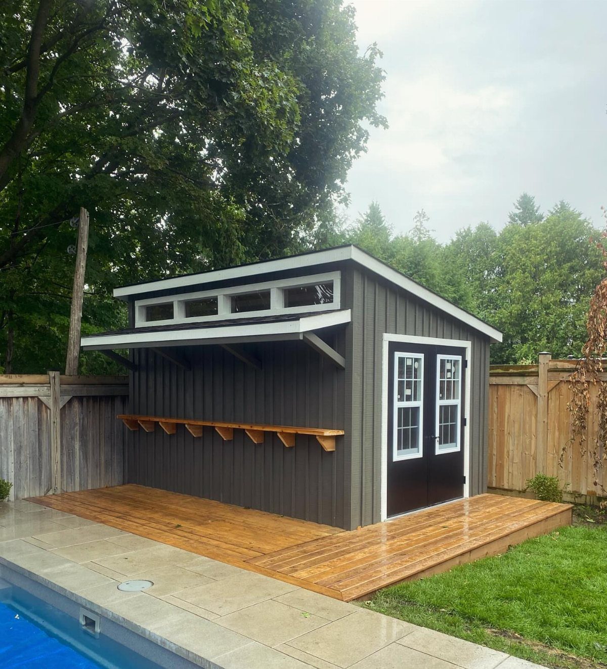 7 Modern Shed Ideas for Compact Yards, Studios & Integrated Outdoor Living 7 A dark grey modern shed with a wooden bar counter and a surrounding wooden deck.
