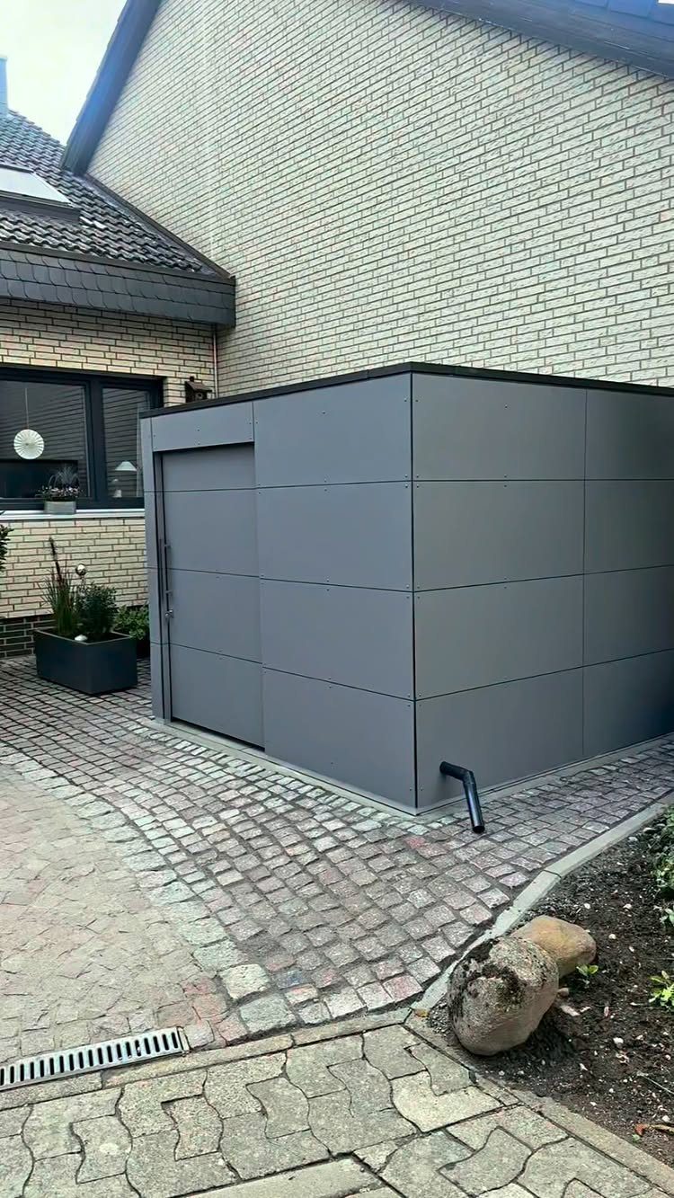7 Modern Shed Ideas for Compact Yards, Studios & Integrated Outdoor Living 6 A modern dark grey paneled shed situated on a cobblestone patio next to a brick house.