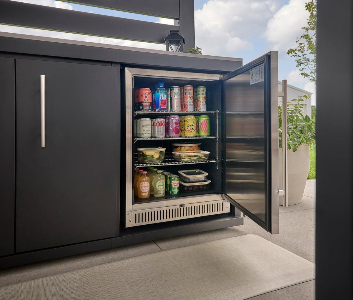 5 Outdoor Kitchen Fridge Ideas for Your Patio Setup 4 An open under-counter refrigerator stocked with drinks and food, built into dark grey outdoor kitchen cabinetry.