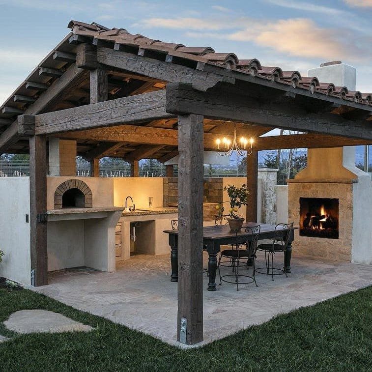 7 Outdoor Patio Kitchen Ideas for Every Backyard Style 1 A rustic wooden pavilion housing an outdoor kitchen with a pizza oven and a stone fireplace.