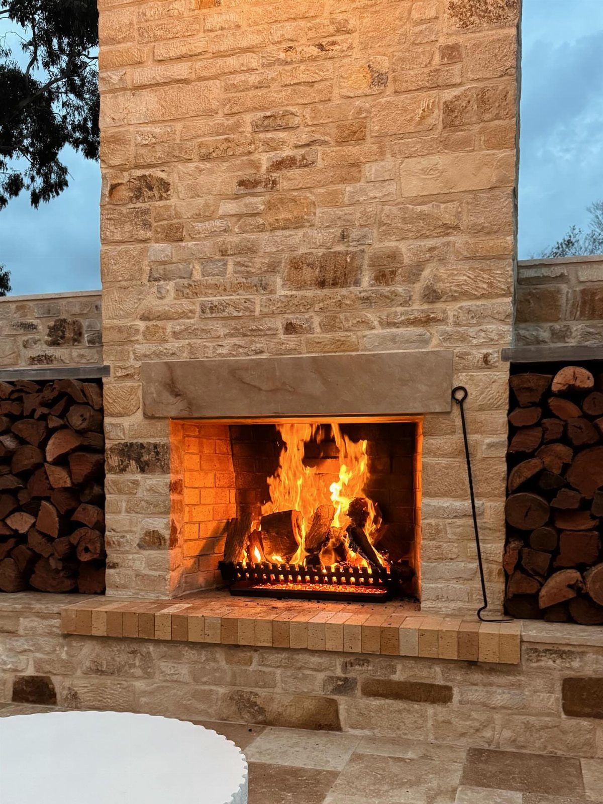 7 Outdoor Patio Kitchen Ideas for Every Backyard Style 5 A large stone outdoor fireplace with a bright fire burning inside and firewood stacked on the sides.