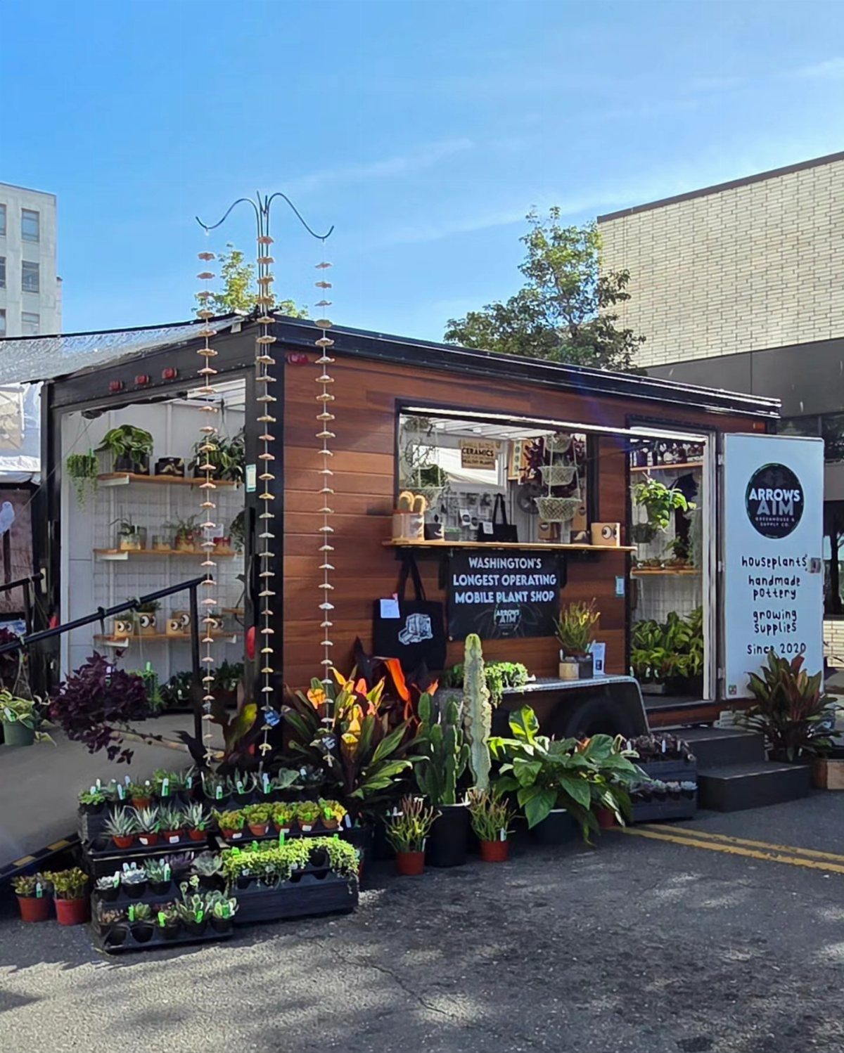 A mobile wooden plant shop trailer displaying numerous potted plants and succulents outdoors.