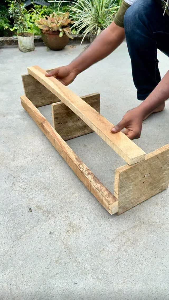 Hands assembling a wooden structure made of light planks on a concrete floor outdoors.