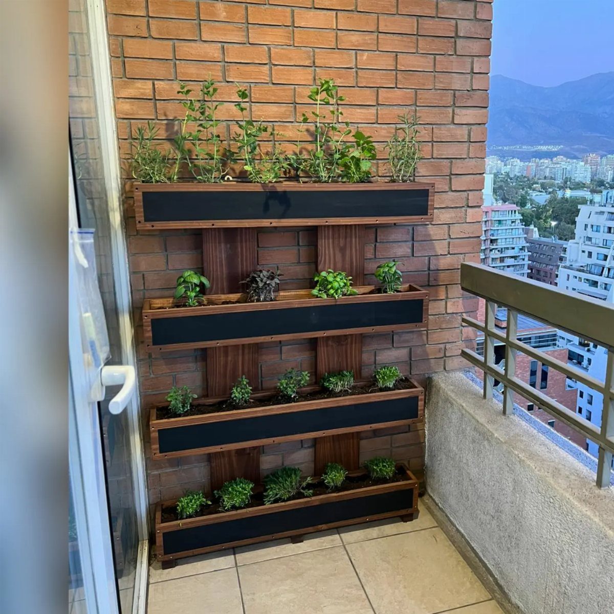 A wooden vertical herb shelf with four black planter boxes mounted on a brick balcony wall.
