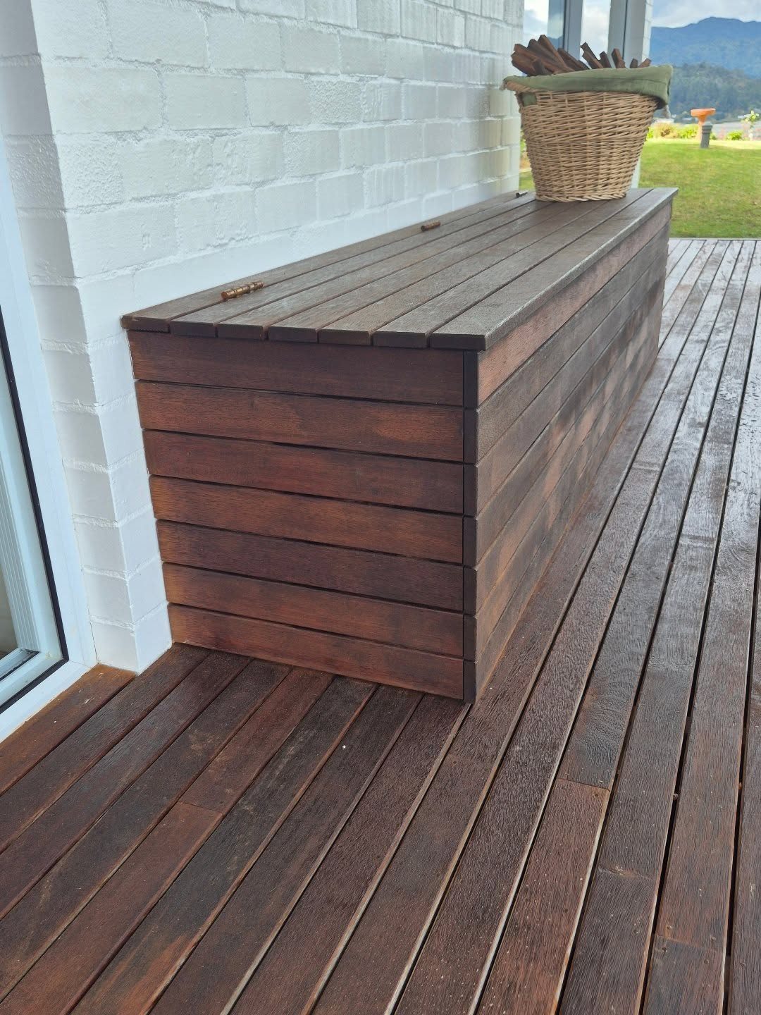 28 Outdoor Wooden Deck Ideas for Any Yard Size 6 A long dark wood storage bench sits on a wooden deck against a white brick wall.