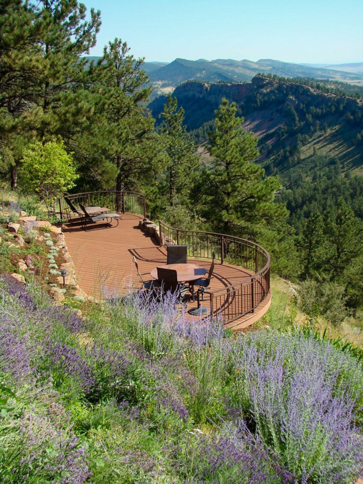 28 Outdoor Wooden Deck Ideas for Any Yard Size 13 Curved wooden deck overlooking a lush green mountain valley with purple wildflowers in the foreground.
