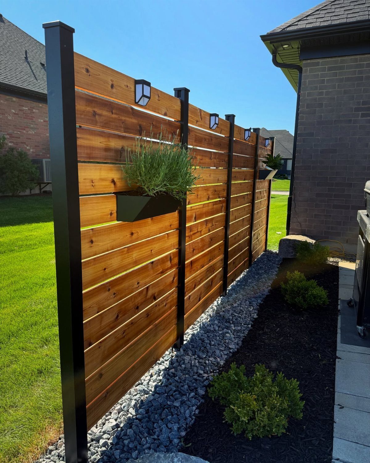 20 Patio and Deck Ideas to Make Your Yard Feel Like a Room 7 Wooden privacy fence with black metal posts and mounted black planter boxes containing green plants.