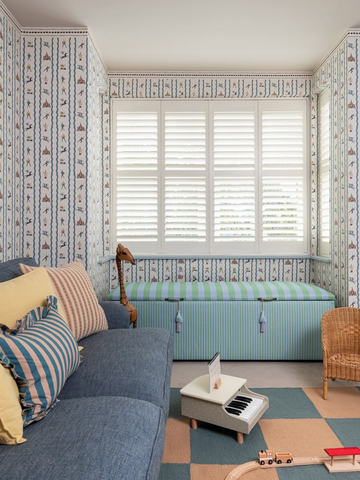 A striped blue and green storage ottoman positioned under a window in a patterned playroom.