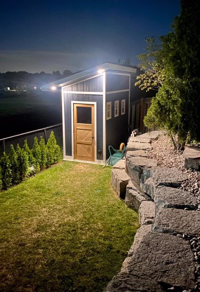 10 Shed Colors Ideas to Paint Your Backyard Storage 9 A navy blue garden shed with white trim and a wooden door at night.