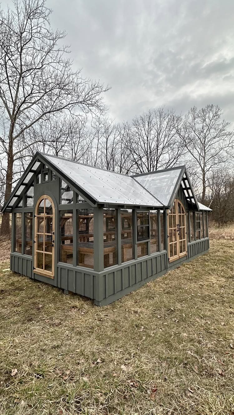 9 Shed Exterior Ideas: Metal, Wood, Recycled, and Garden Styles 5 A grey wooden greenhouse-style shed with large glass windows and arched doors in a field.