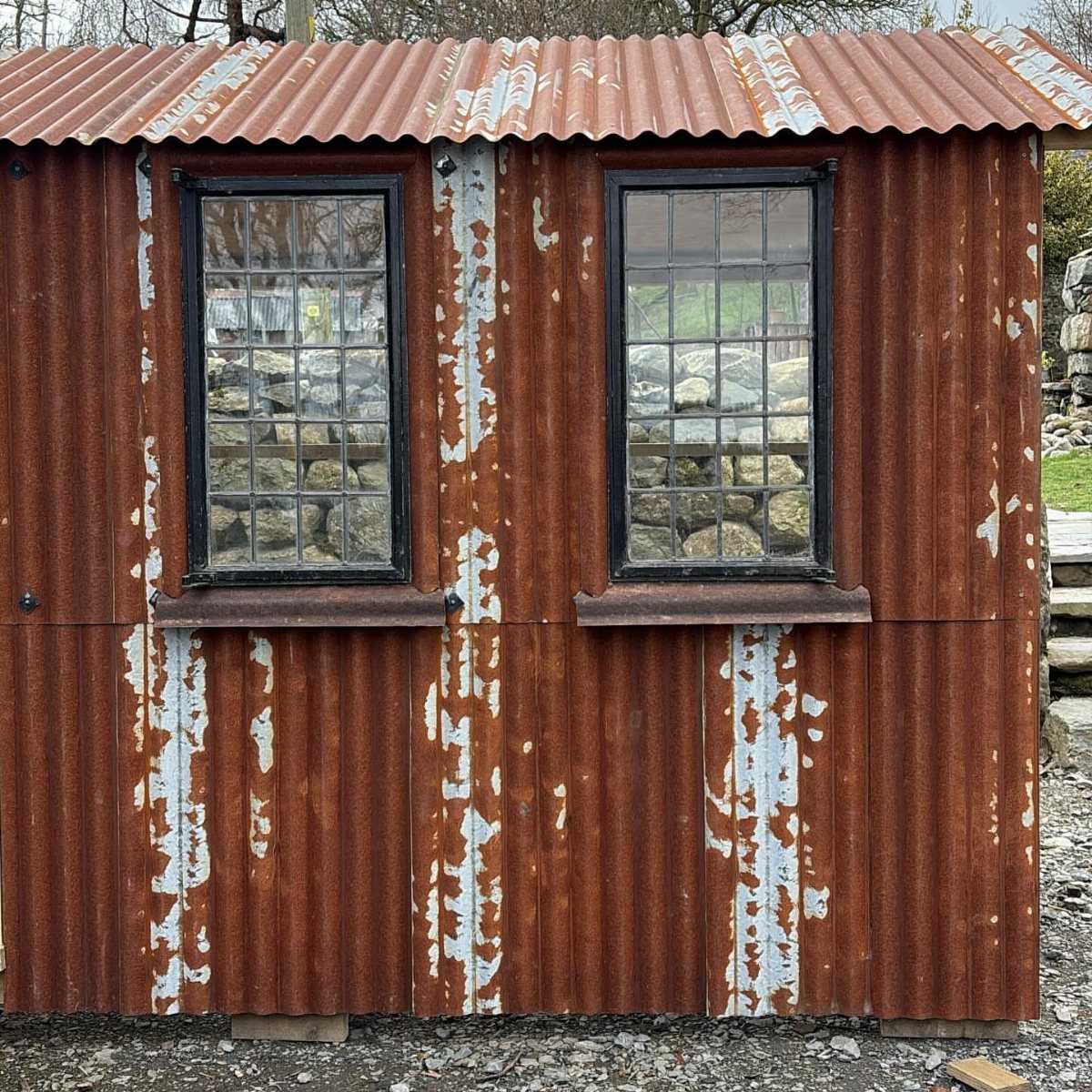 9 Shed Exterior Ideas: Metal, Wood, Recycled, and Garden Styles 3 A rustic shed featuring heavily rusted corrugated metal siding and two small black-framed windows.
