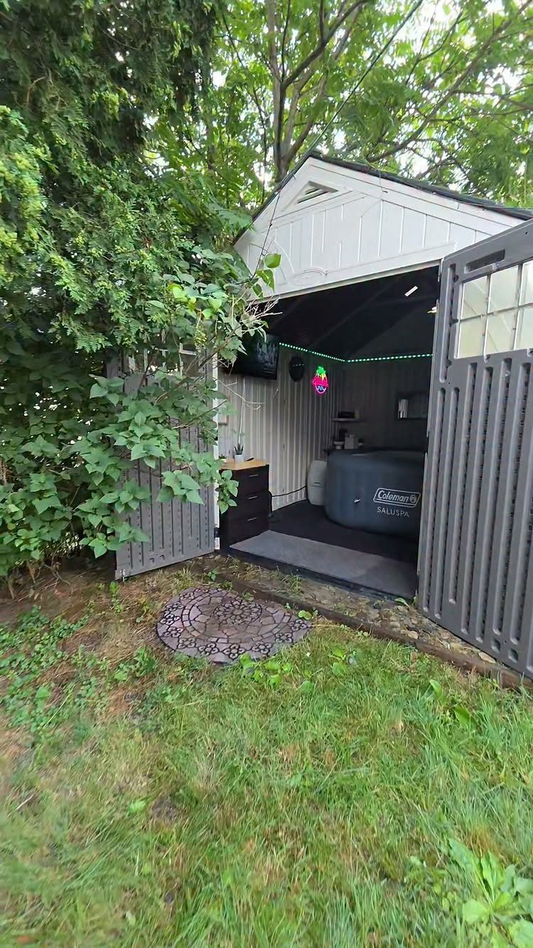 Grey wooden shed with open doors containing a hot tub and string lights in a backyard.