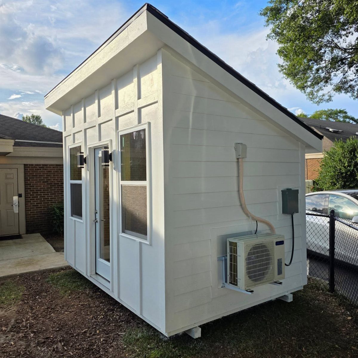 White wooden studio shed with an external air conditioning unit mounted on the side wall.