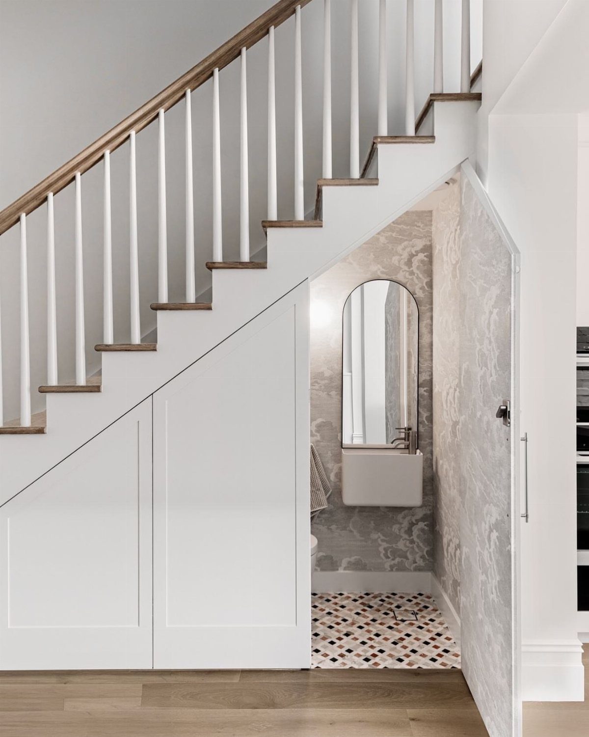 18 Under Stair Storage Design Modern Ideas for Every Home 10 A small powder room with a sink and arched mirror located under a white staircase.