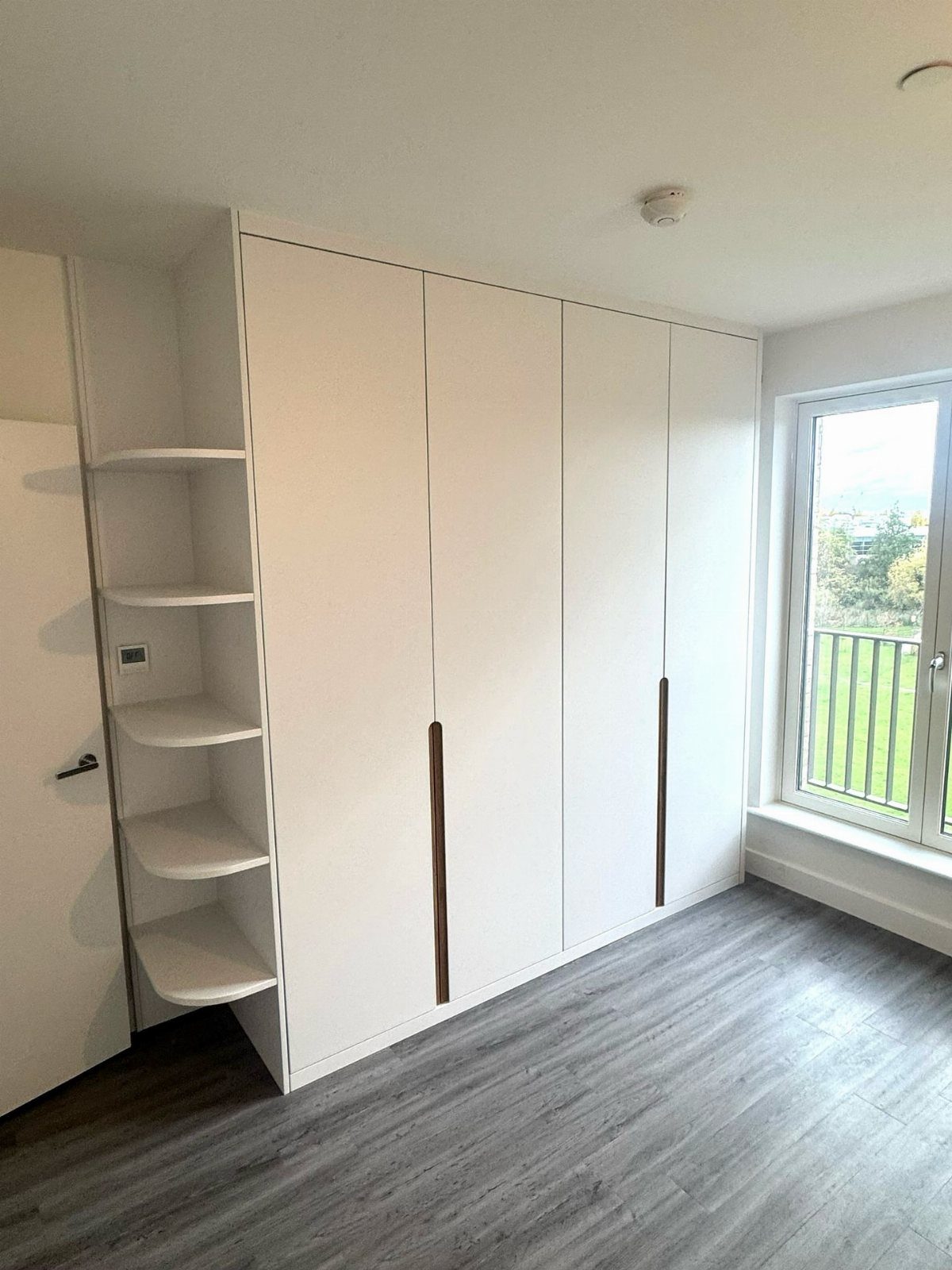 White floor-to-ceiling wardrobe with wooden handles and open corner shelving in a bright room.