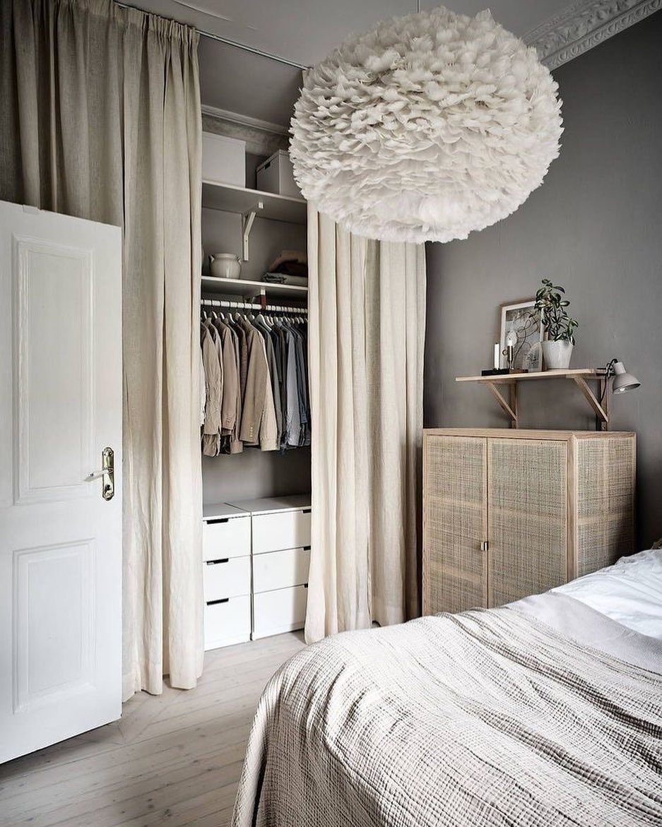 Open wardrobe with hanging clothes and white drawers behind beige sliding curtains in a grey bedroom.