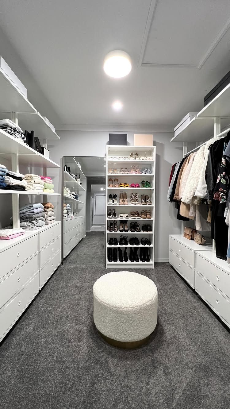 White modular shelving and hanging clothes in a bright walk-in closet with a white ottoman.