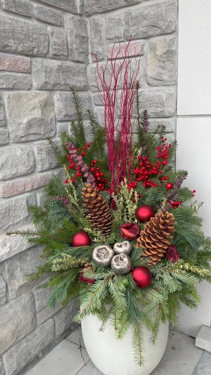 7 Winter Garden Room Ideas: Plants, Protection and Whimsy 4 Evergreen branches with red berries, pinecones, and ornaments in a white planter against a stone wall.