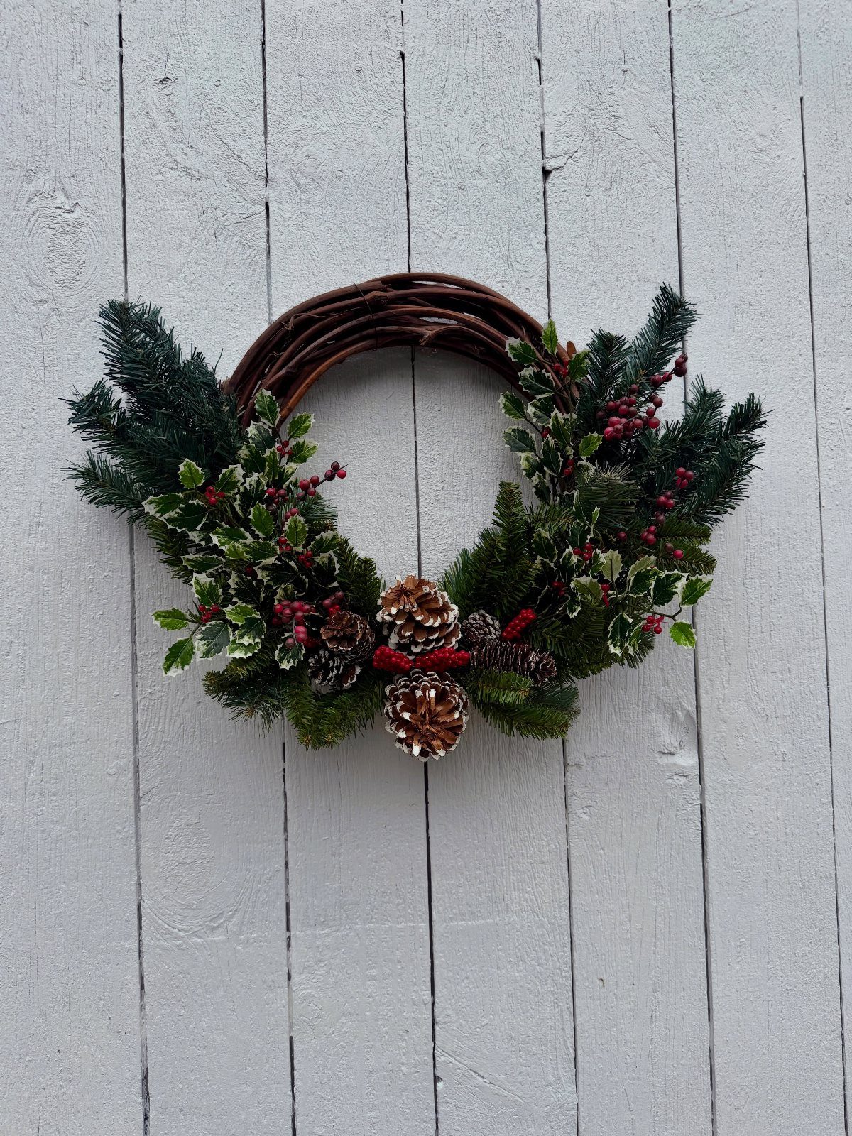 A grapevine wreath with evergreen branches, holly, red berries, and pinecones on white wood.