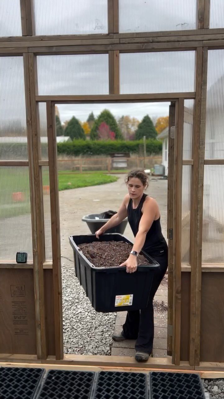 A woman carrying a large black storage bin filled with soil through a greenhouse door.